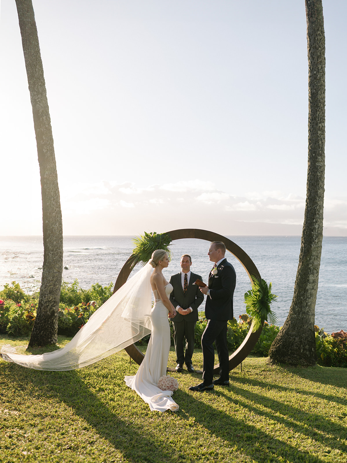 Wide view of the couple standing under a circular arch with palm trees and the ocean at Merriman’s Kapalua.
