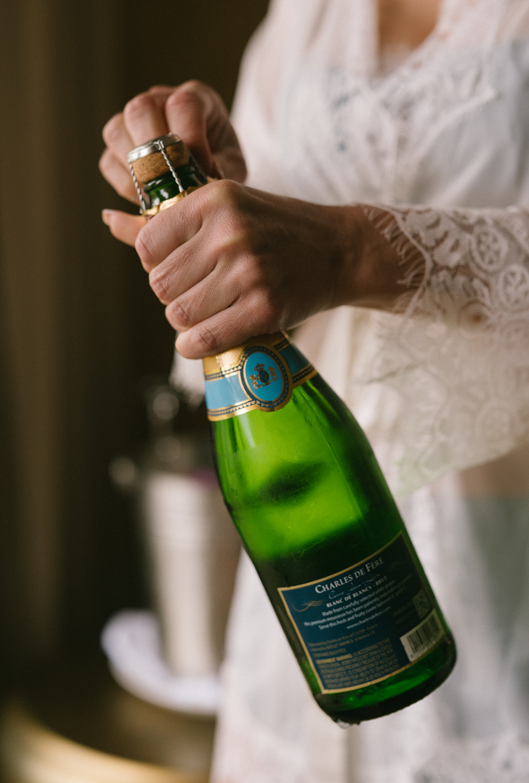 Bride opening a bottle of champagne while wearing a lace robe during wedding preparations.
