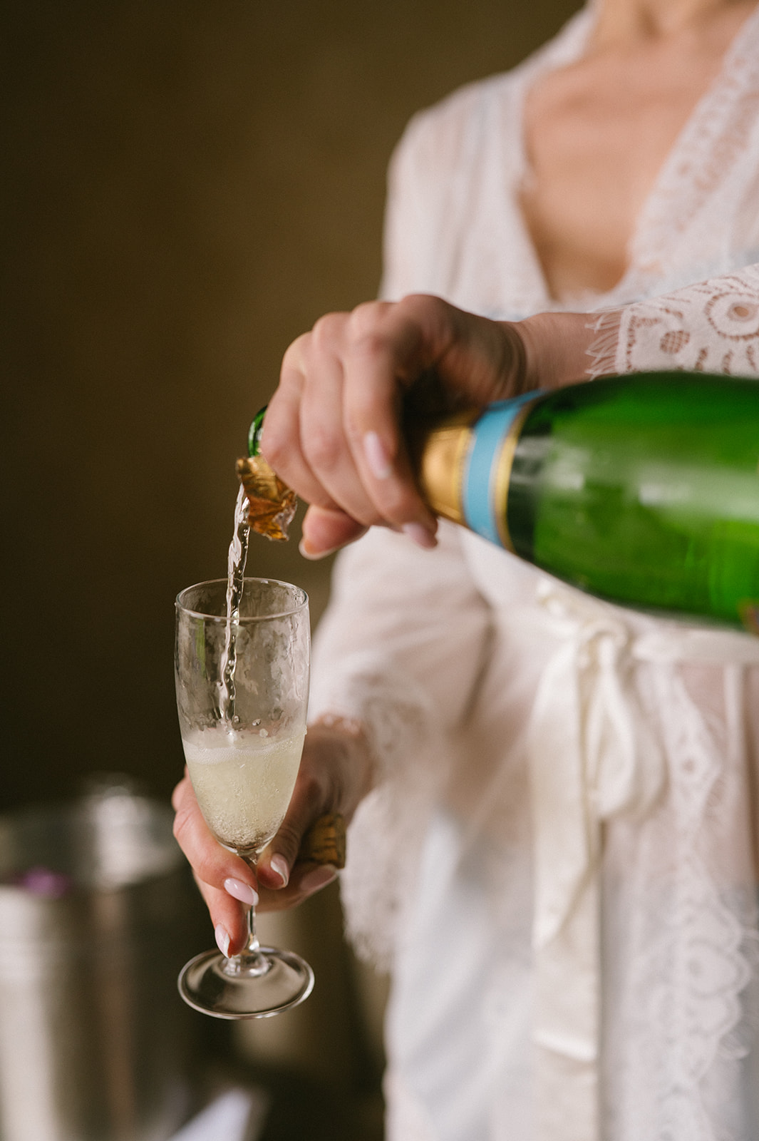 Bride pouring champagne into a flute while wearing a lace robe in soft window light.
