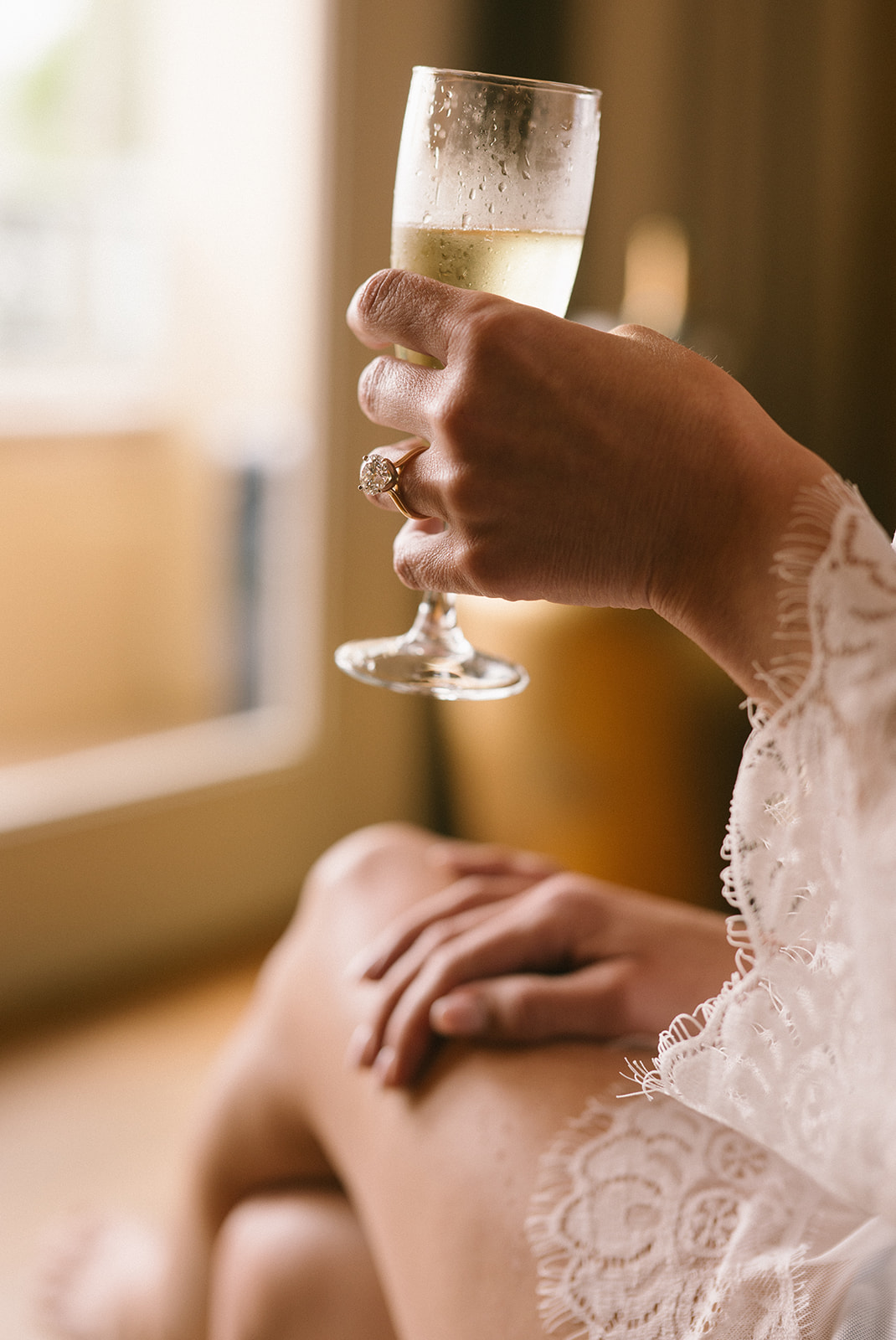 A bride’s hand, adorned with a sparkling engagement ring, holds a champagne glass during wedding morning preparations.