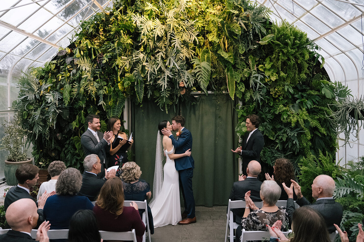 Taylor and Patrick sharing their first kiss as a married couple in front of a greenery wall.