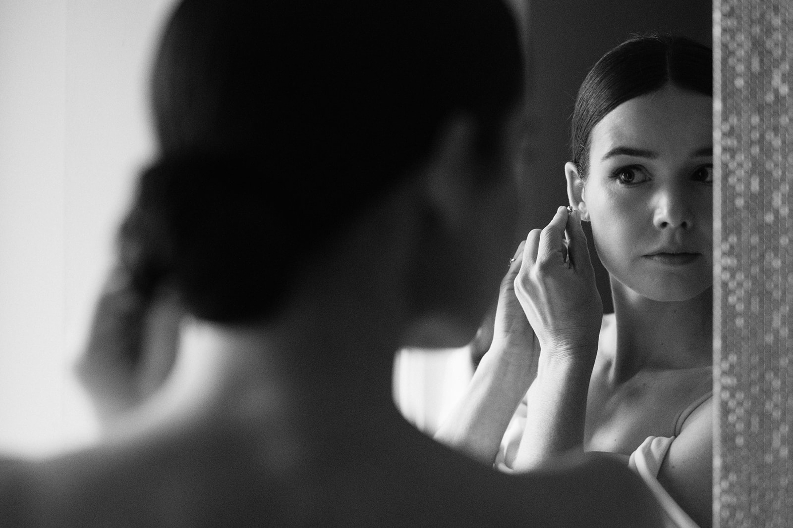 A black-and-white shot of a bride adjusting her earrings, reflected in the mirror, with an elegant and focused expression.