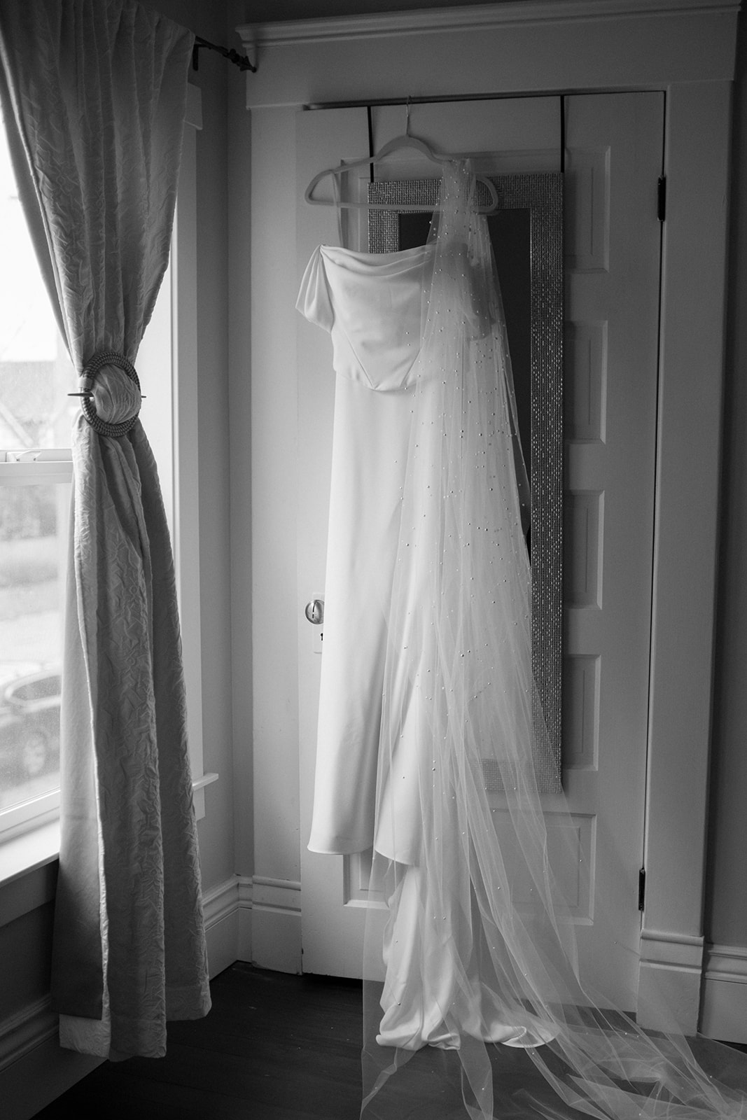 A wedding dress with a flowing veil hangs on a door beside a bright window, ready for the bride.