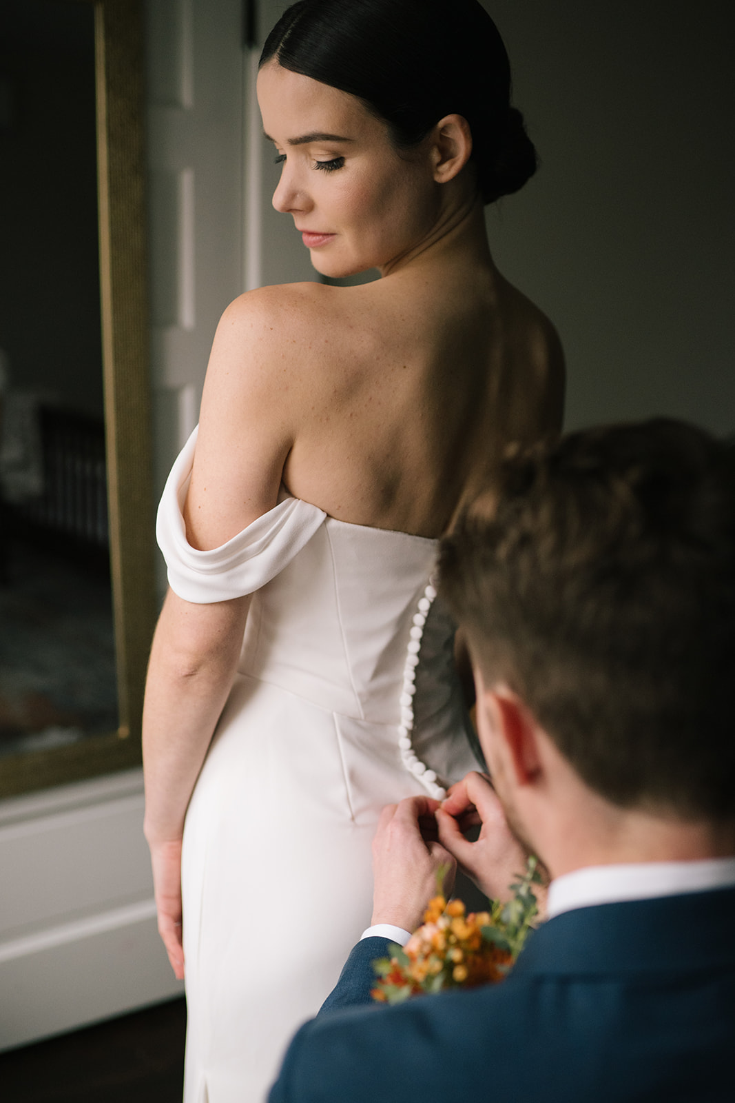 A groom carefully buttons the bride's elegant wedding gown, framed in soft natural light.