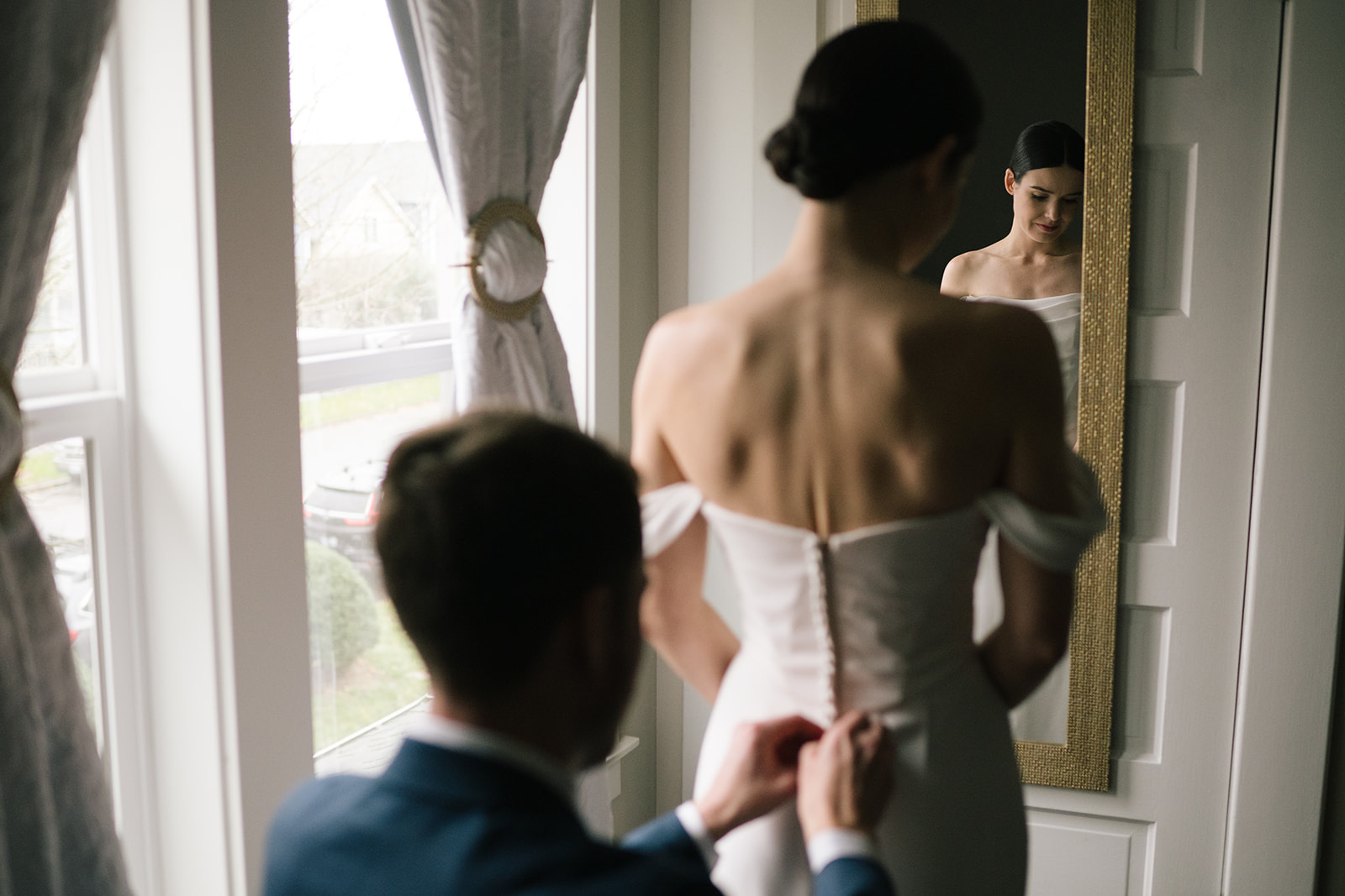 A bride in her wedding dress gazes into a mirror as the groom fastens the buttons on her gown.