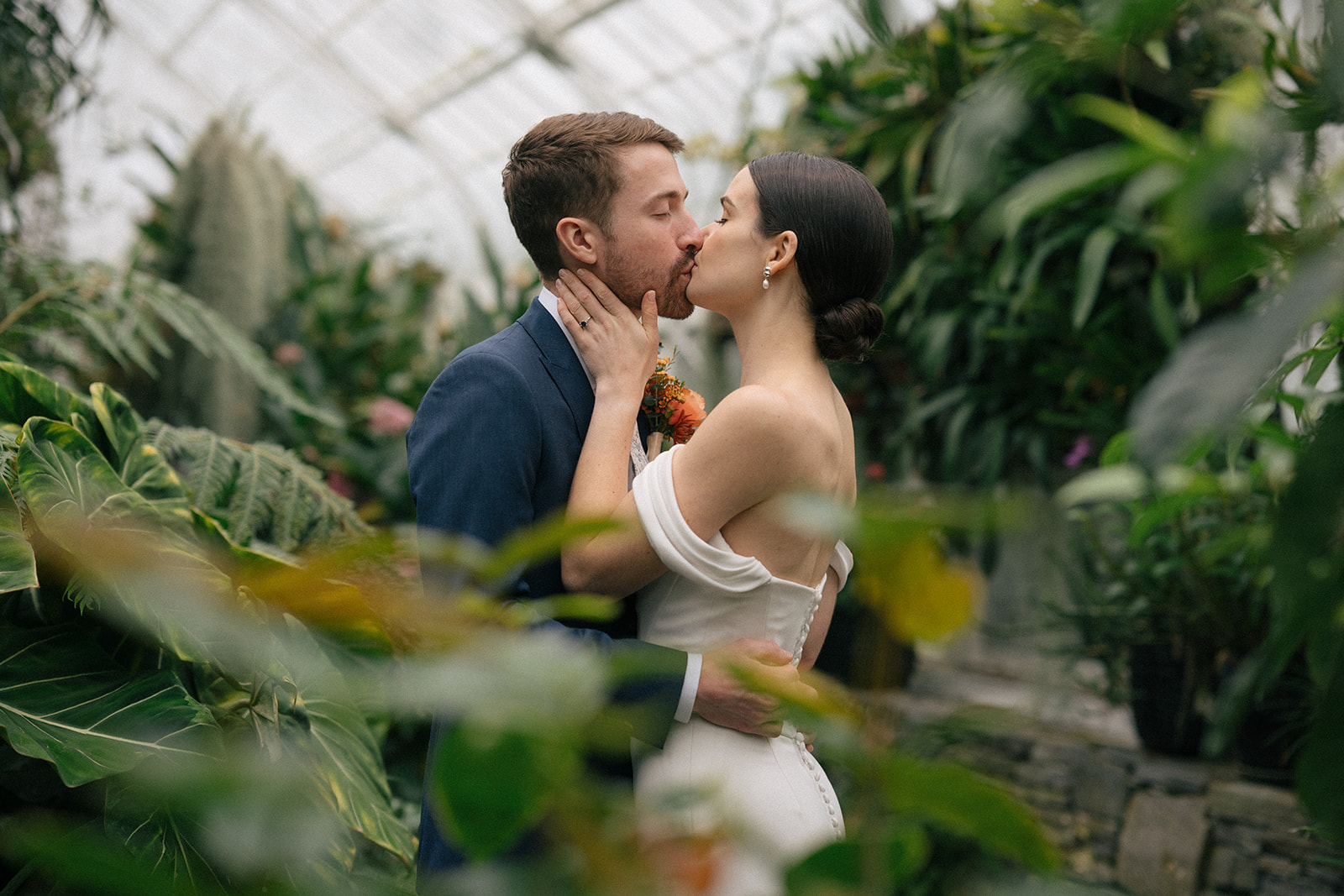 Taylor and Patrick sharing a kiss framed by vibrant plants inside the Seymour Conservatory.