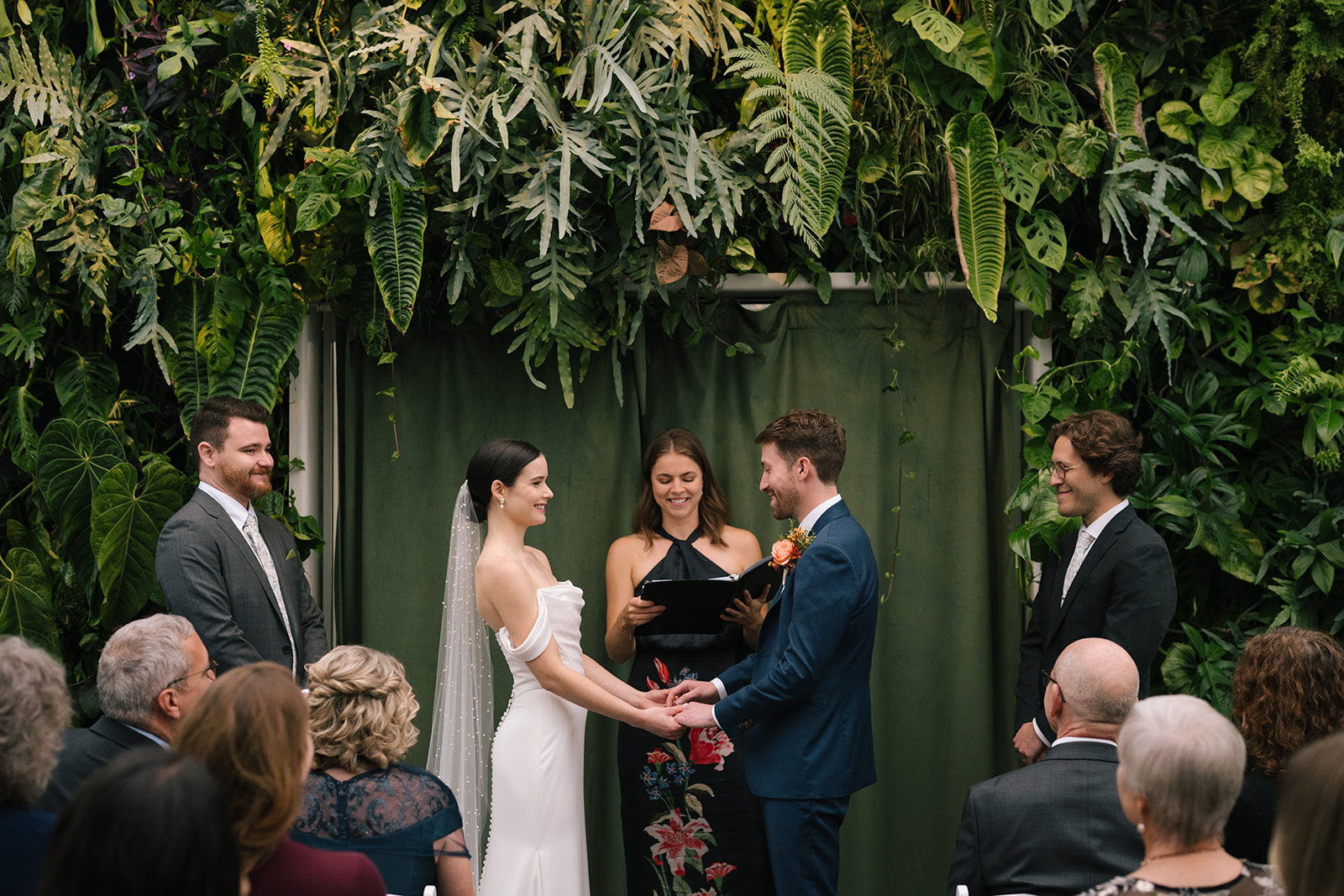 Taylor and Patrick holding hands during their ceremony in front of a vibrant wall of greenery.