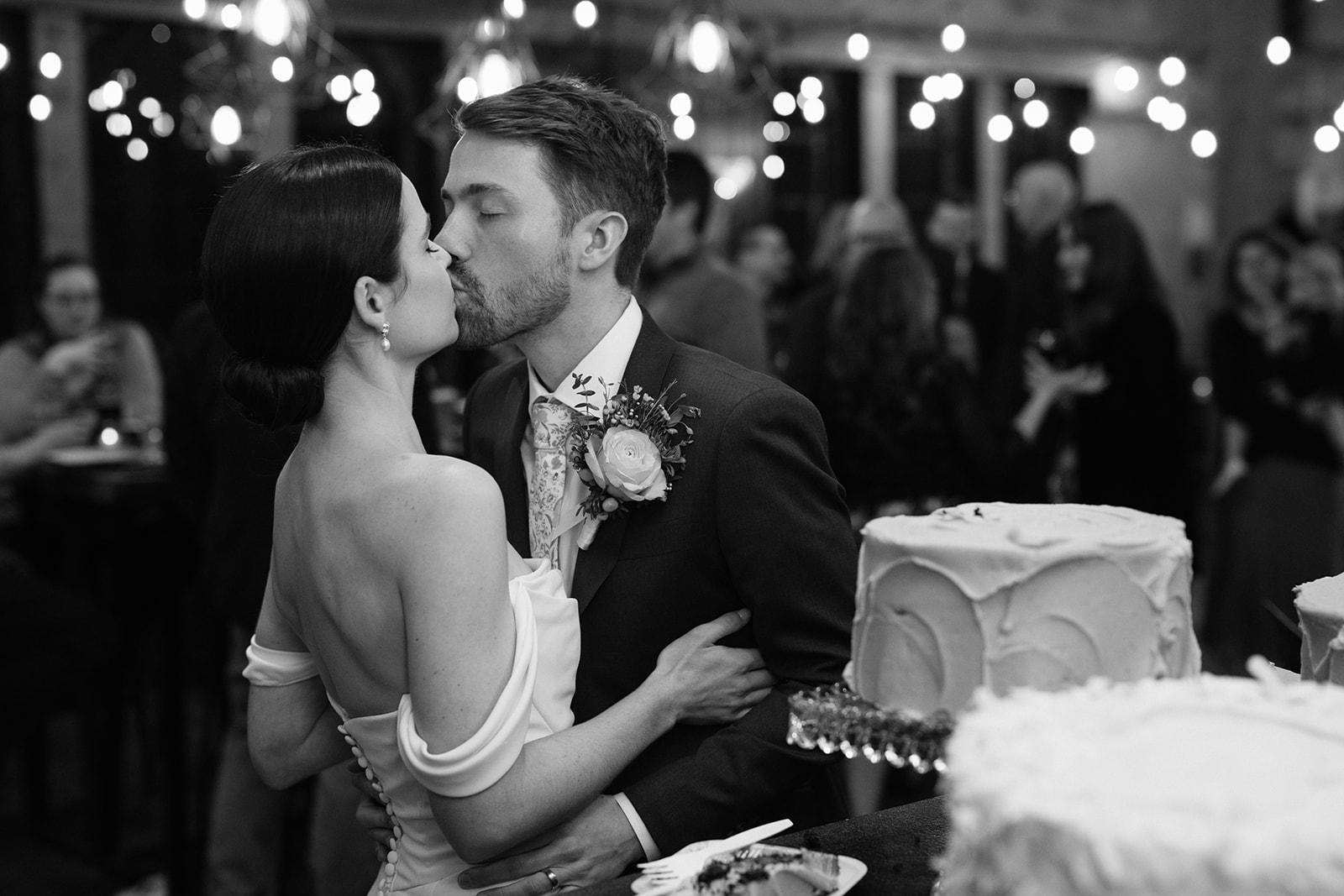 Bride and groom share a kiss during their wedding reception by the cake table.