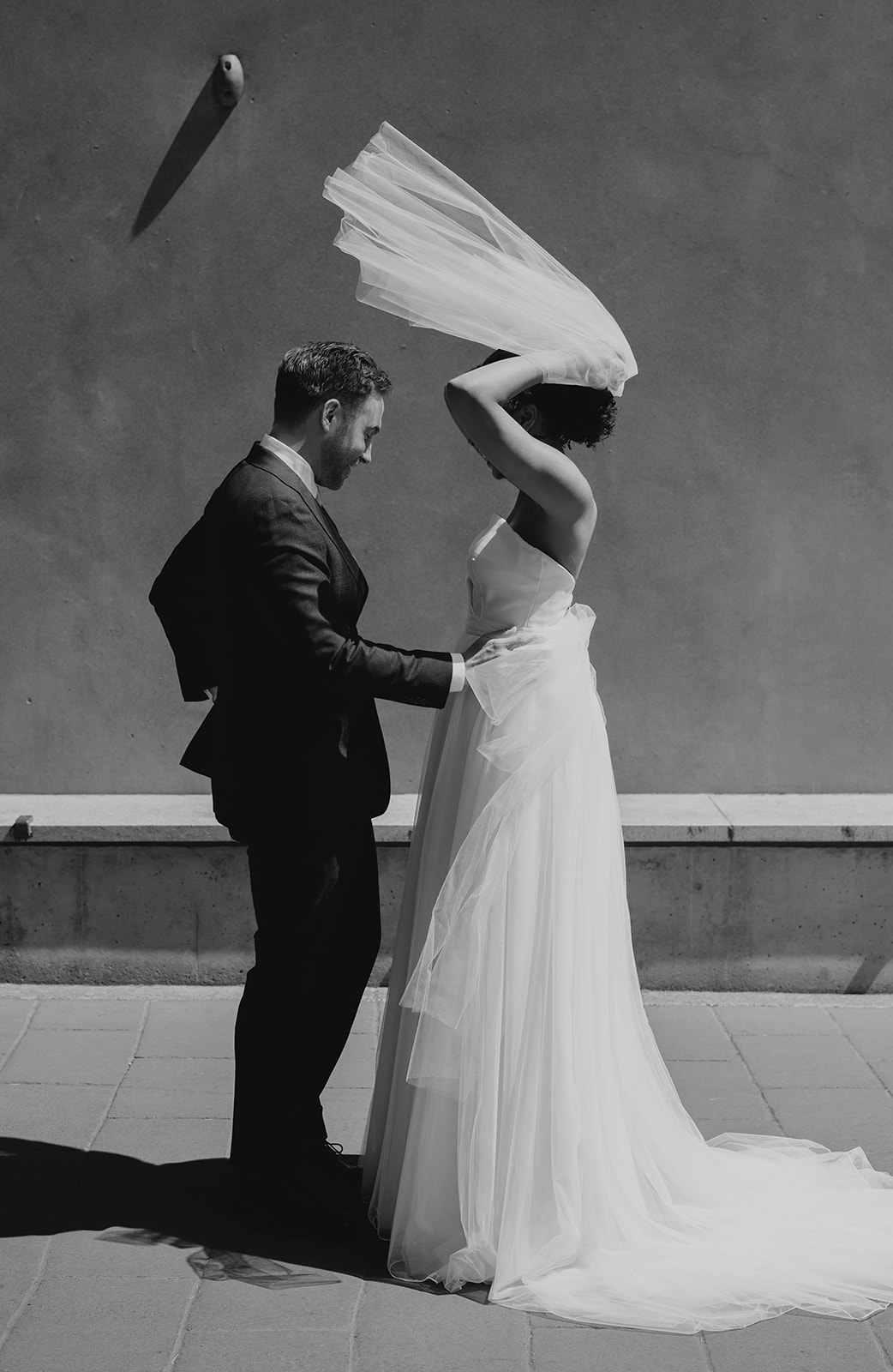 Bride lifting her veil as groom adjusts her dress during a black and white wedding portrait