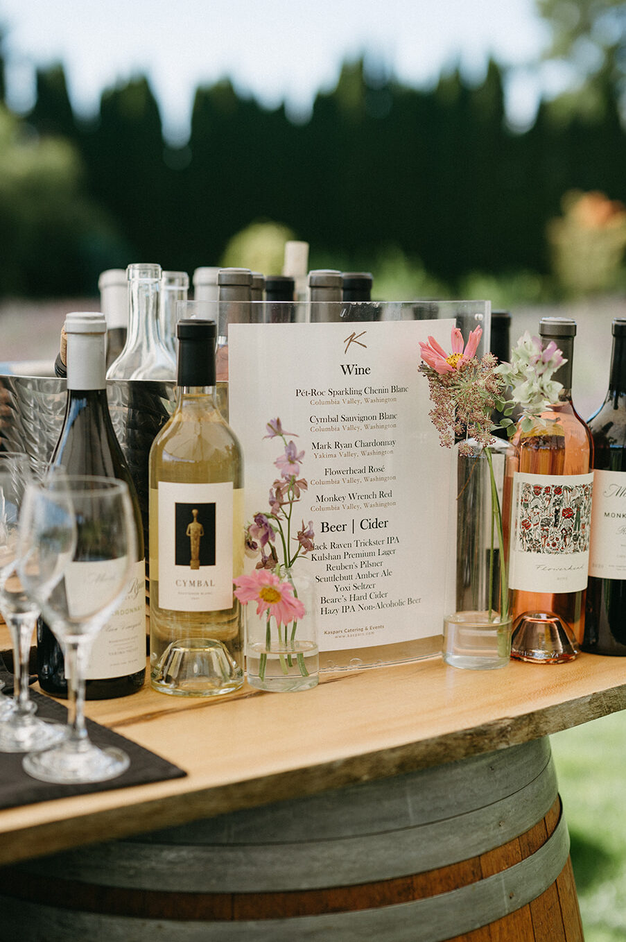 Wine and cider bar setup with floral accents and a printed drink menu at wedding reception.
