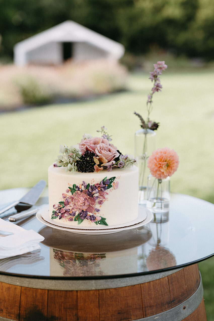 Single-tier white cake decorated with purple flowers and fresh blooms on a wine barrel.
