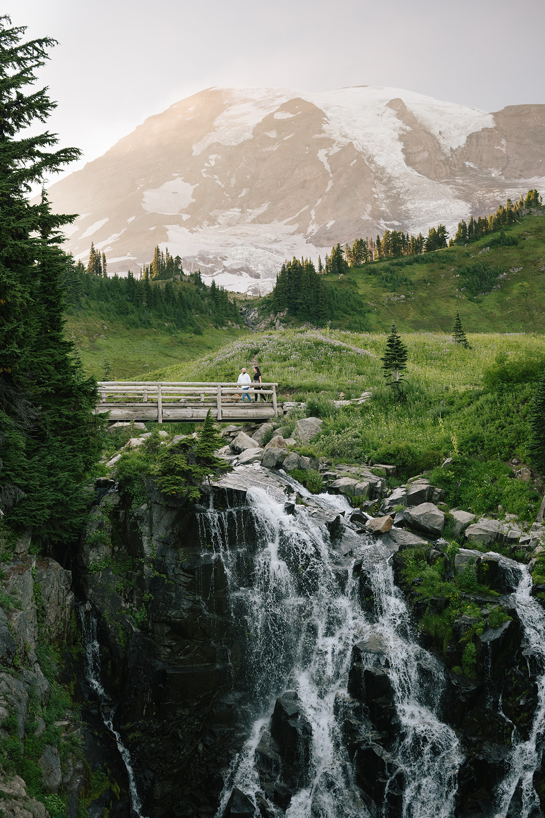 Wide landscape of the Myrtle Falls footbridge with a newly engaged couple after their surprise Mount Rainier proposal