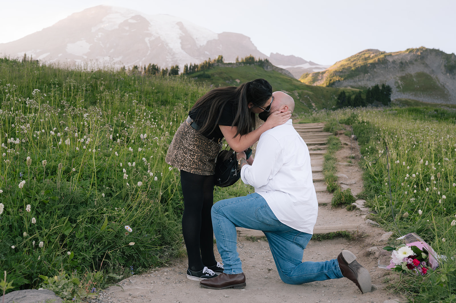 Bride-to-be leans in to kiss her fiancé on the forehead as he kneels on the trail during a surprise Mount Rainier proposal, marker bouquet visible on the ground