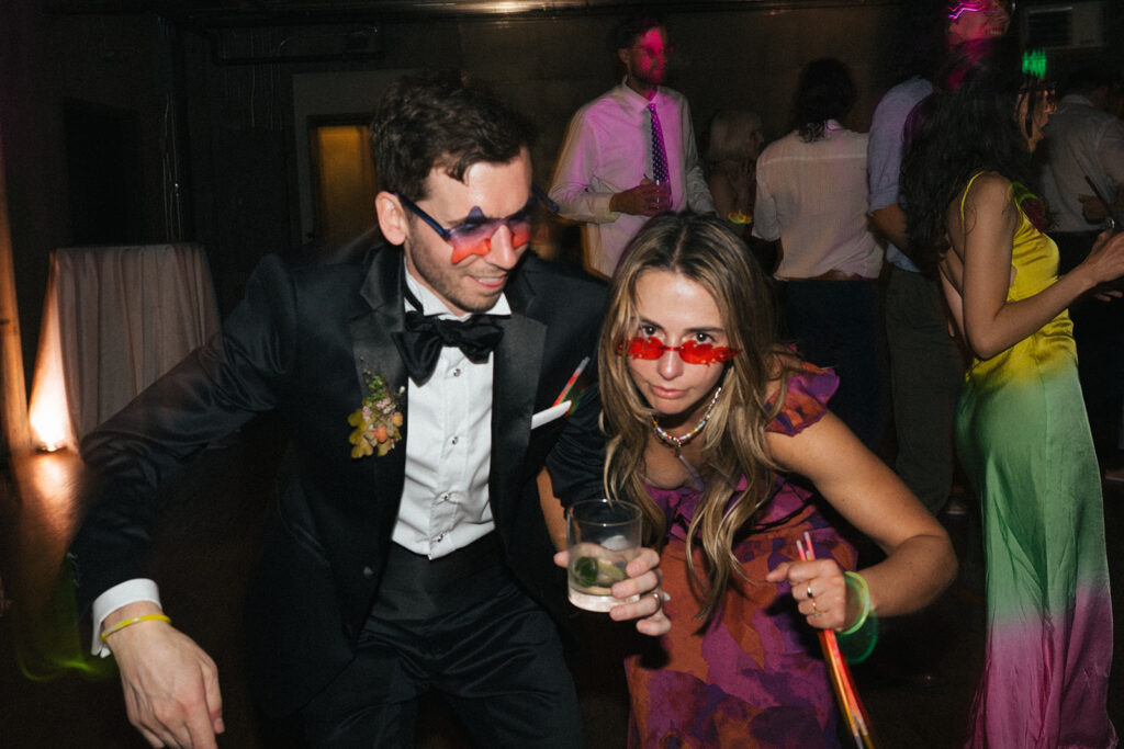 Groom and a guest wearing fun sunglasses and holding drinks while dancing at the Fremont Foundry wedding
