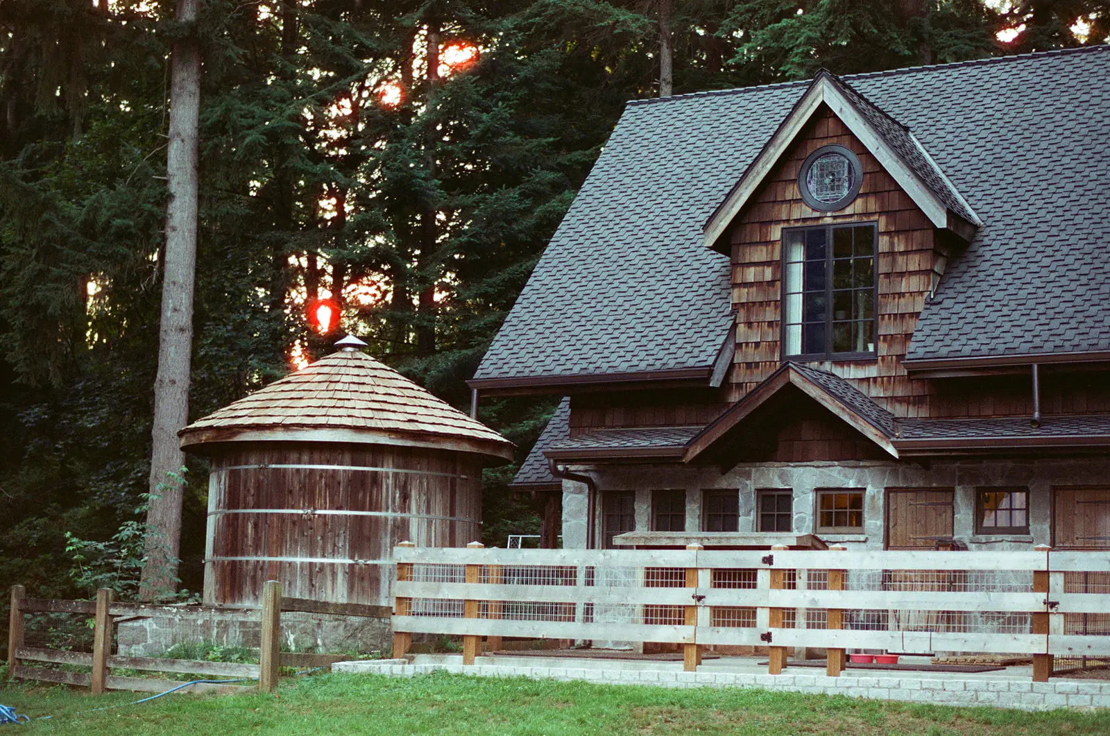 The cedar-shingled cottage and round wooden silo at Bella Luna Farms in Snohomish, Washington, photographed on Candido ISO 800 film as the sun sets behind the trees.
