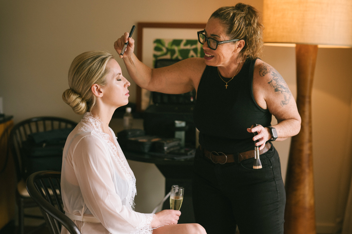 Bride having her makeup applied by a makeup artist while seated in her room at the Four Seasons Maui, holding a glass of champagne.
