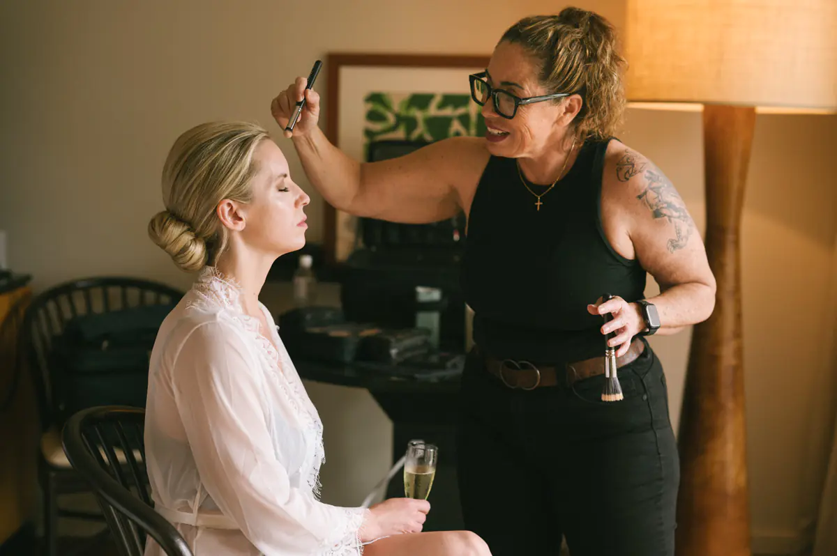 Bride having her makeup applied by a makeup artist while seated in her room at the Four Seasons Maui, holding a glass of champagne.
