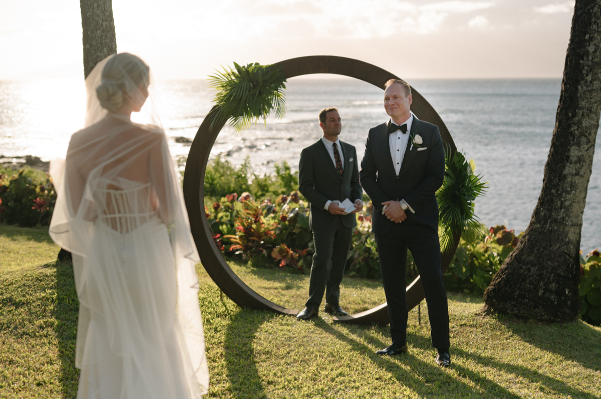 Bride approaching the groom and officiant beneath a circular arch with the ocean behind them at Merriman’s Kapalua.
