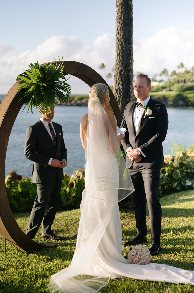 Groom reading his vows to the bride beneath a tropical arch overlooking the ocean in Kapalua.
