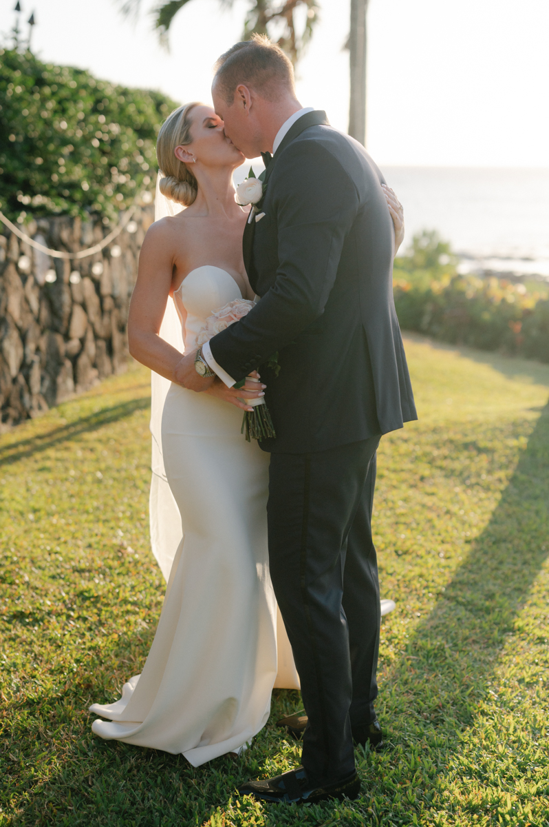 Bride and groom kissing in soft sunset light with the ocean in the background at Merriman’s Kapalua.
