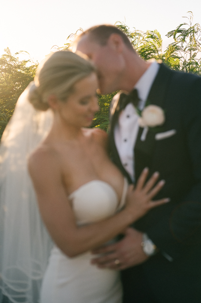 Soft-focus portrait of the bride and groom holding each other in warm sunset light at Merriman’s Kapalua.
