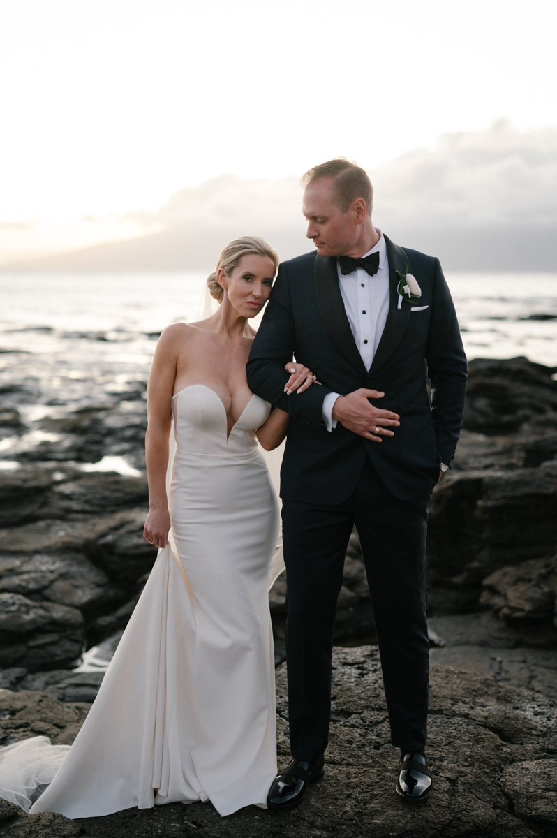 Bride and groom walking arm in arm across the lava rocks with the ocean behind them in Kapalua, Maui.
