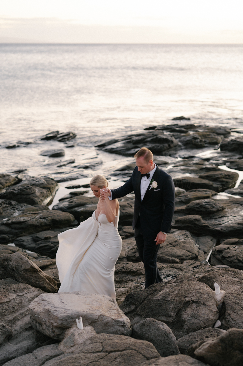 Bride and groom carefully walking together across the lava rocks along the Kapalua coastline at sunset.
