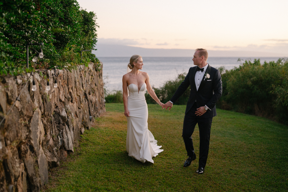Bride and groom walking hand in hand along the lawn at Merriman’s Kapalua with the ocean at sunset.
