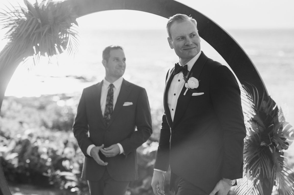 Black and white photo of the groom standing beneath a circular tropical arch overlooking the ocean at Merriman’s Kapalua before the bride arrives.
