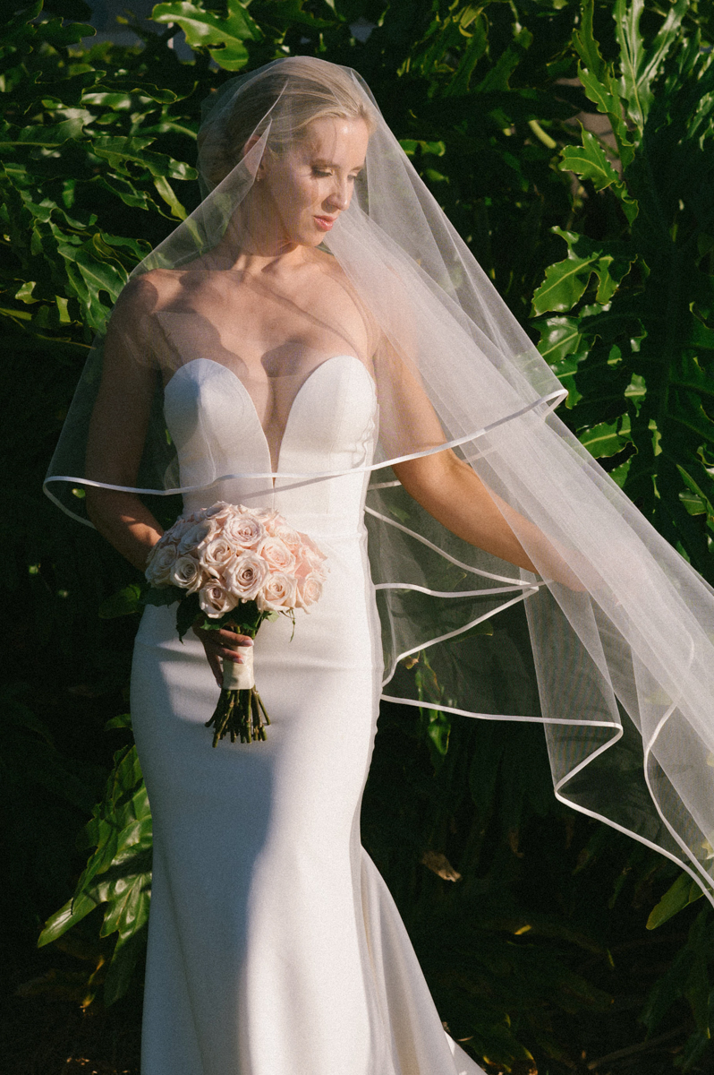 Bride holding her bouquet as her veil drapes over her shoulders in soft golden light in Kapalua.
