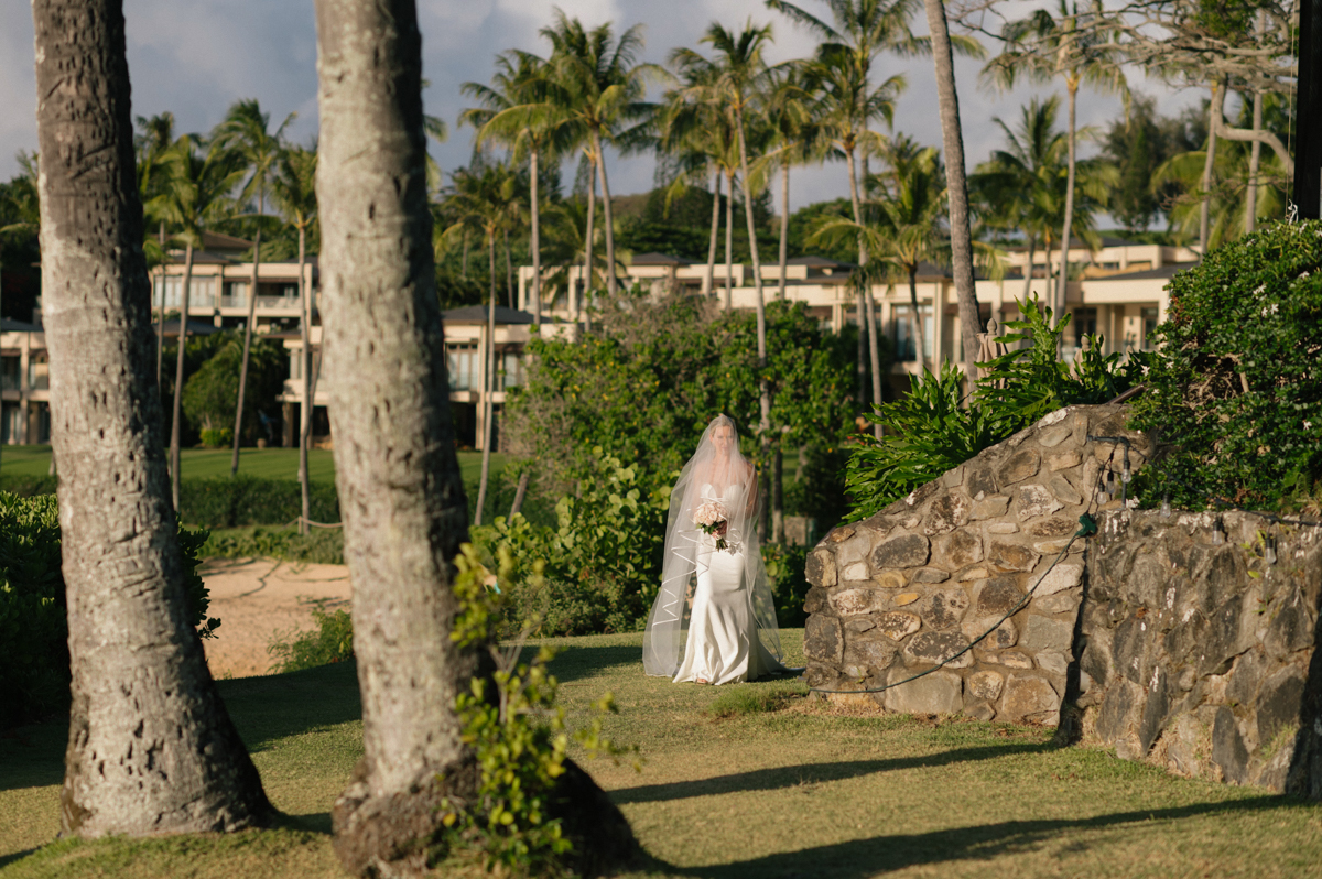 Bride walking through tropical gardens toward the ceremony site at Merriman’s Kapalua with her veil flowing behind her.
