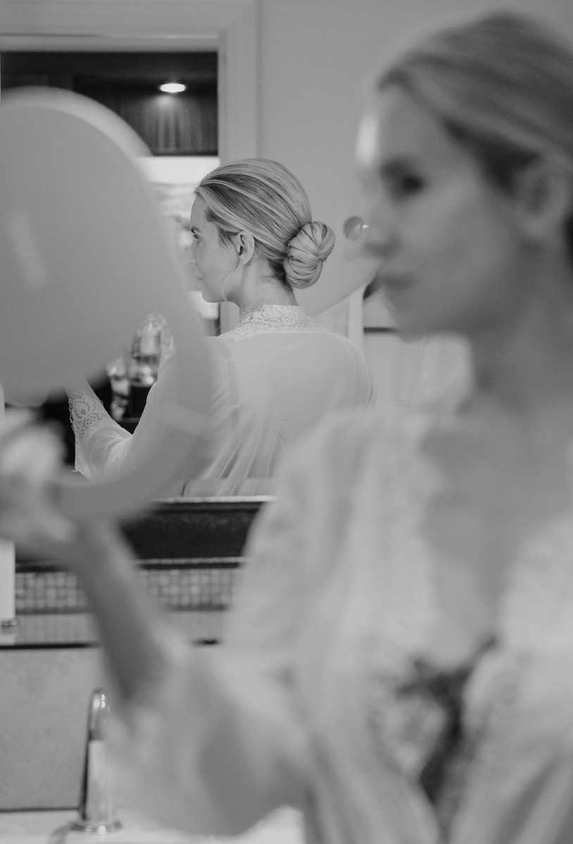 Black and white image of the bride holding a mirror, showing her reflection and low bun hairstyle.
