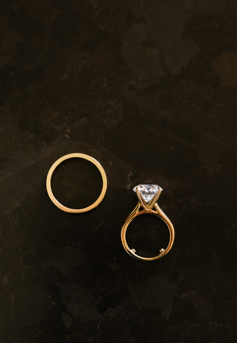Gold wedding band and diamond engagement ring photographed on a dark surface.
