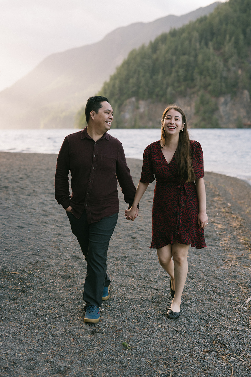 Laughing couple walking hand in hand along the rocky shoreline of Lake Crescent with forested mountains rising behind them, one of the best places to elope on the Washington coast