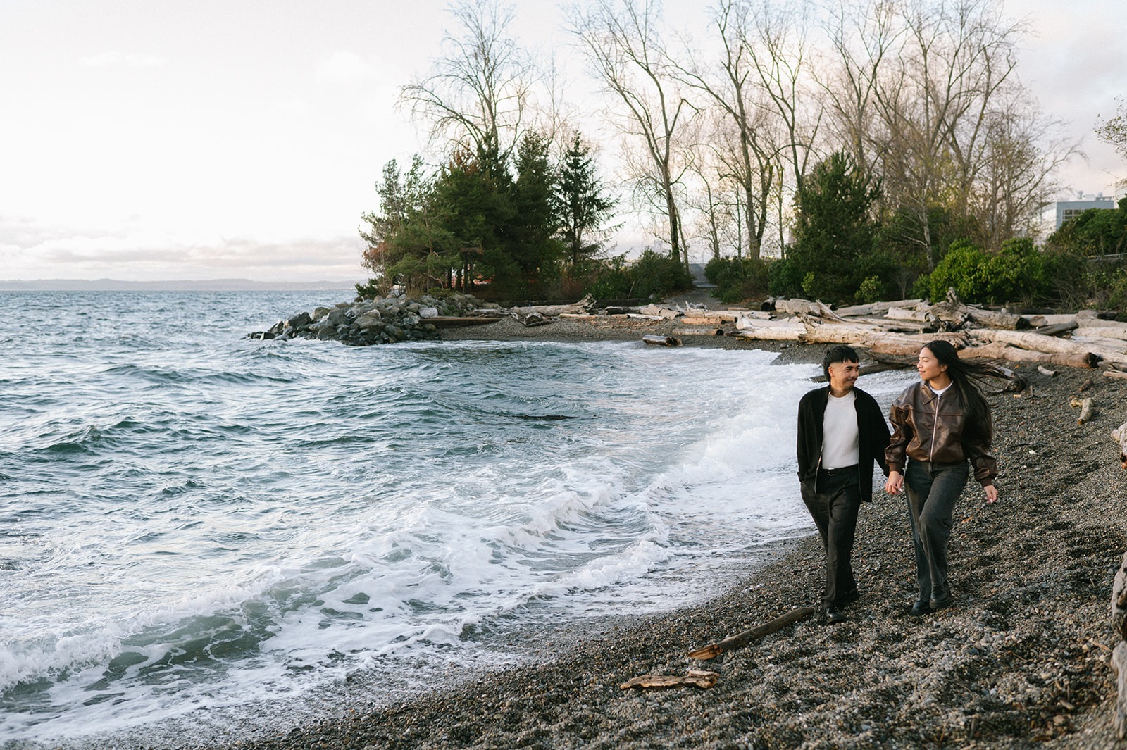 Couple walks hand in hand along the pebble shore of Pocket Beach in Seattle before a surprise proposal