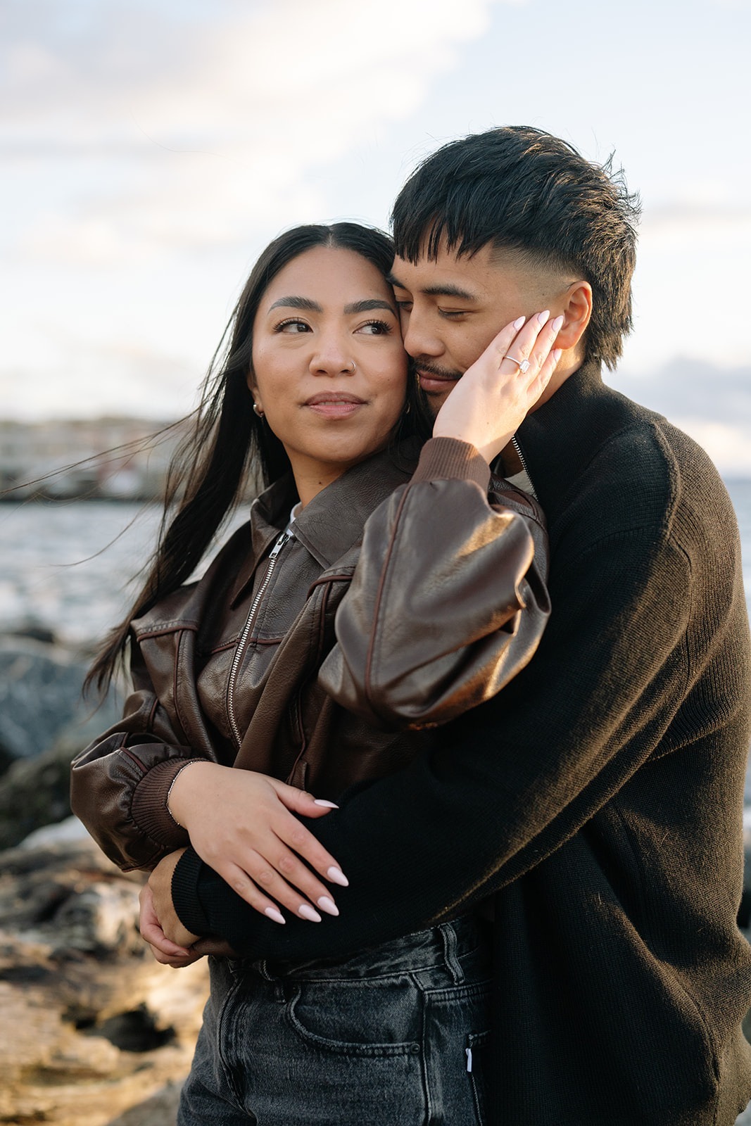 Bride-to-be tenderly touches her fiancé's face, engagement ring visible, at Pocket Beach in Seattle
