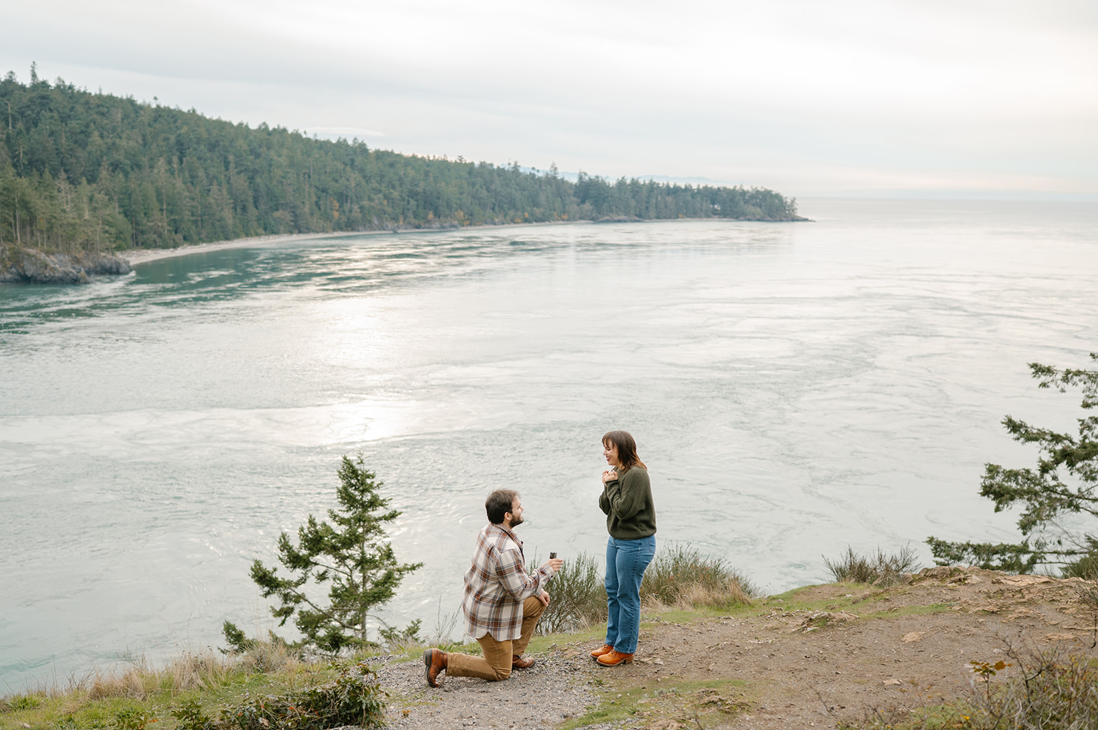 Jonathan kneels on the cliffside as Ashley stands in front of him, the water stretching out behind them.