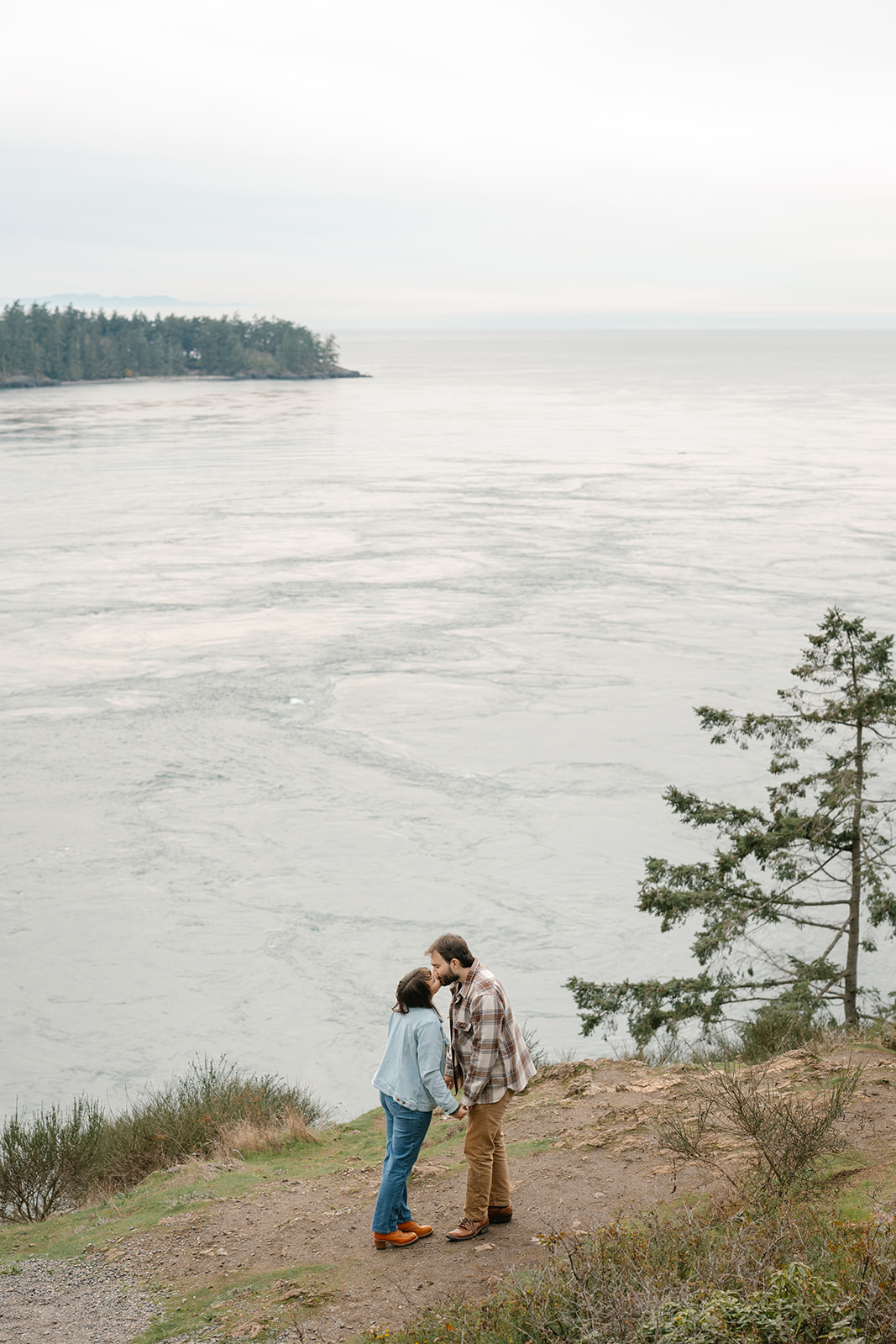 Jonathan and Ashley stand together on a cliff kissing by the water at Deception Pass, small against the vast landscape.