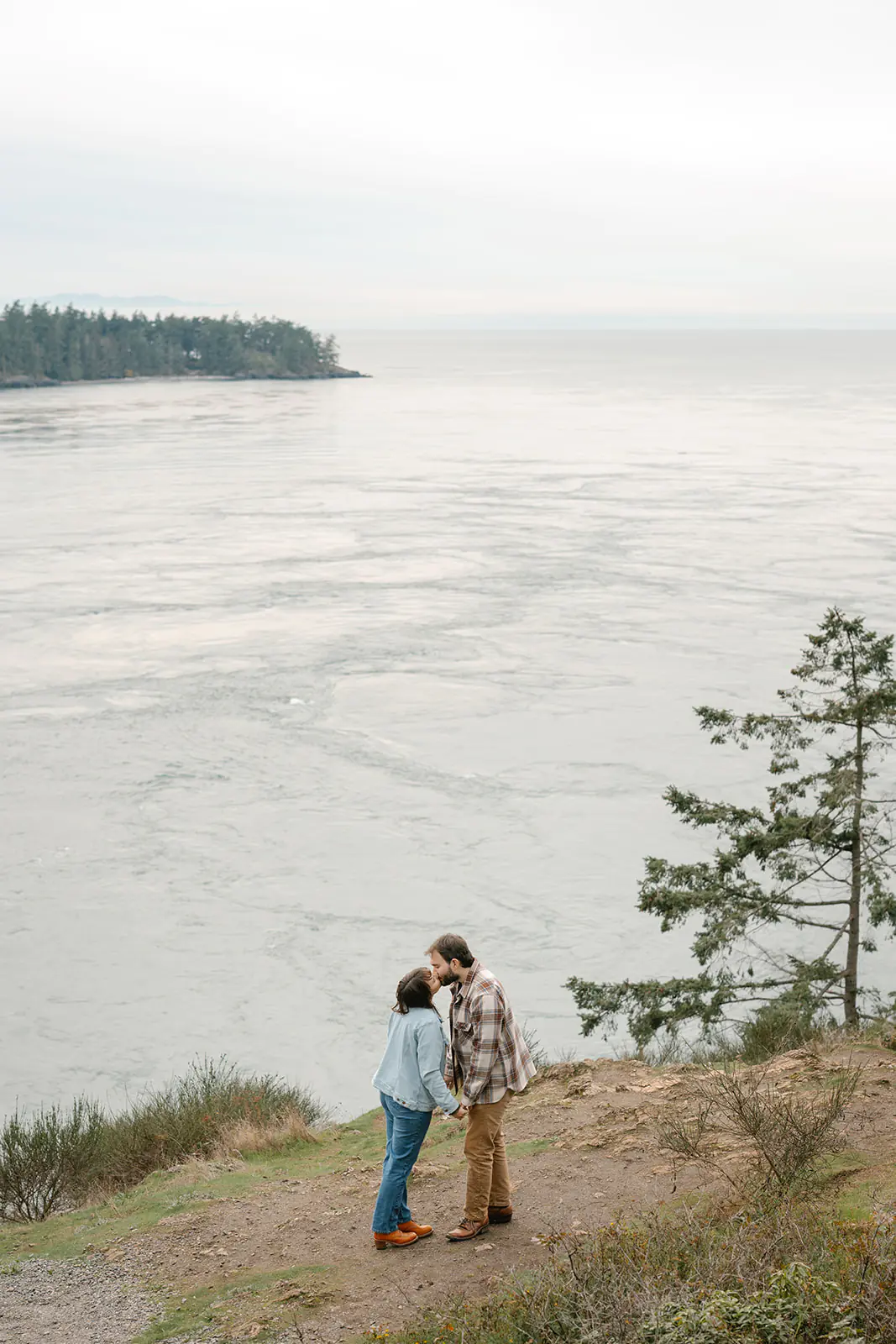 Jonathan and Ashley stand together on a cliff kissing by the water at Deception Pass, small against the vast landscape.