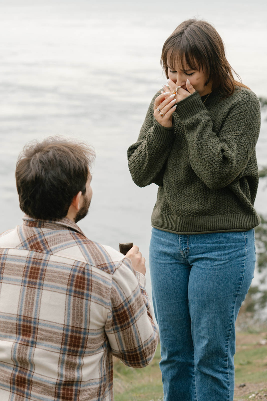Jonathan kneels in front of Ashley holding the ring box as she stands in stunned happiness.
