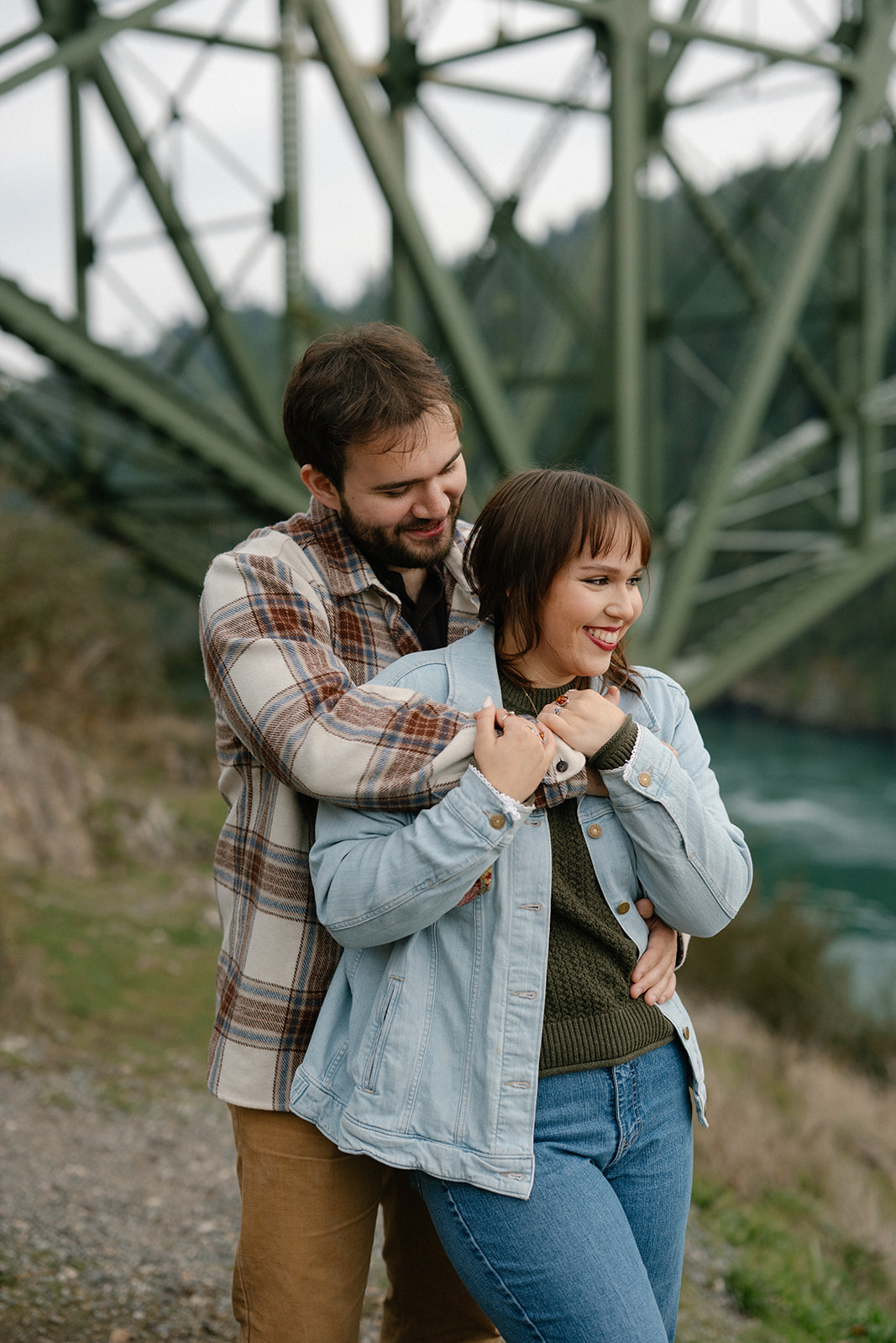 Jonathan hugs Ashley from behind with the Deception Pass Bridge in the background.
