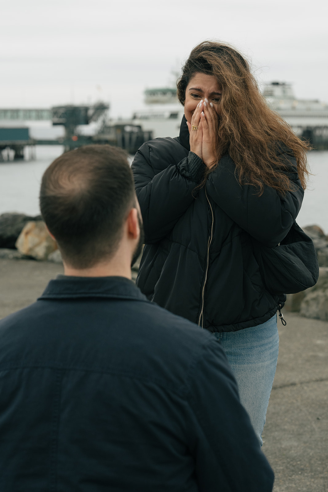 Aysha covers her face in happy shock as Brad proposes by the water.