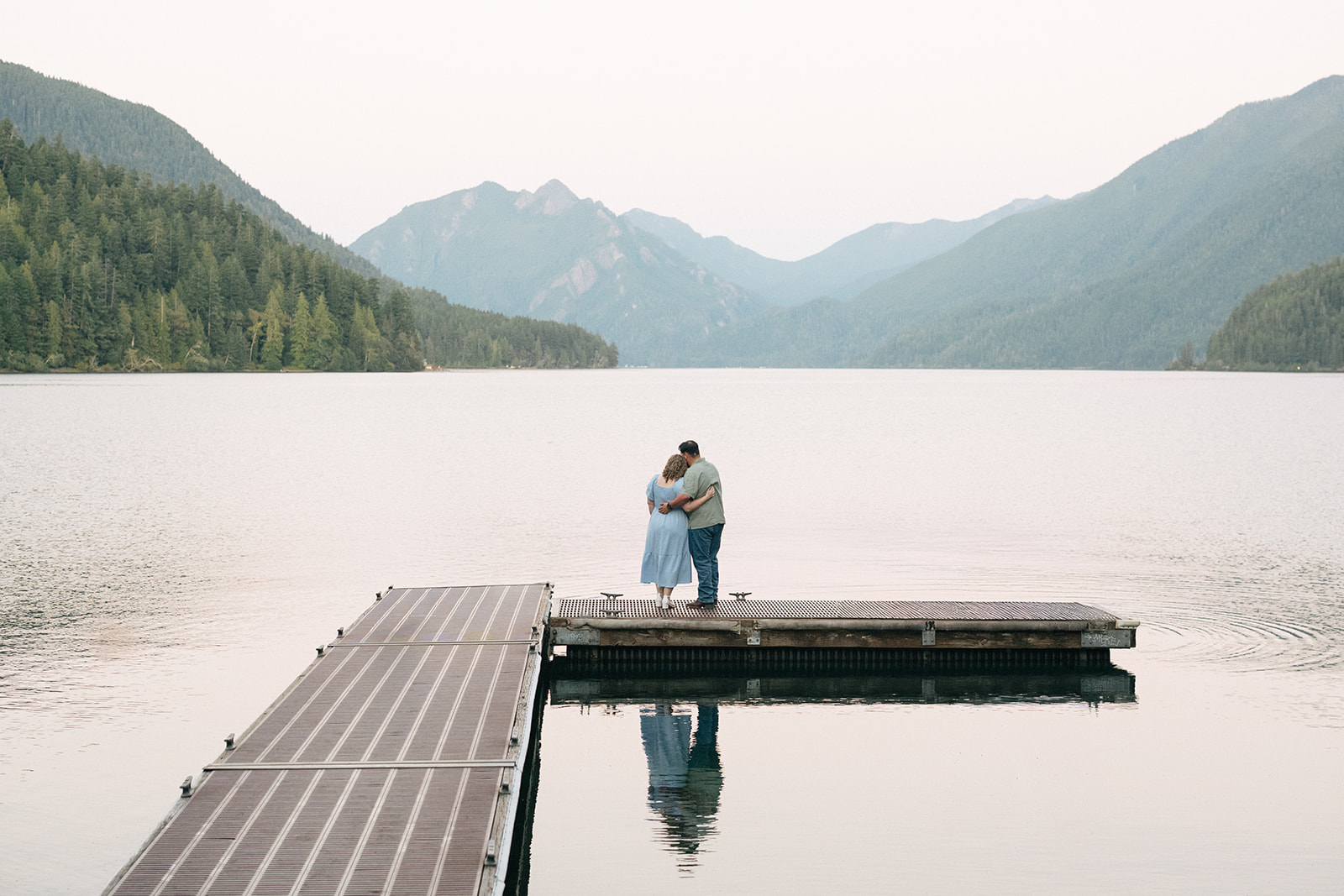 Couple standing together on a dock at Lake Crescent looking out at calm water and forested mountains, Olympic National Park Washington