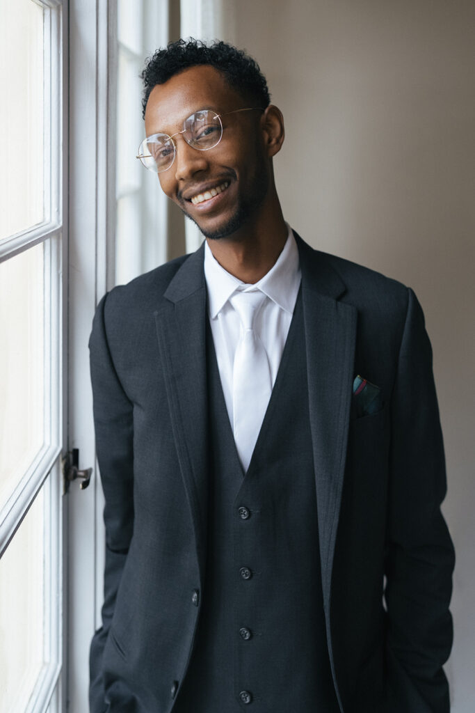 Groom in black suit standing by window light at Lairmont Manor before wedding ceremony