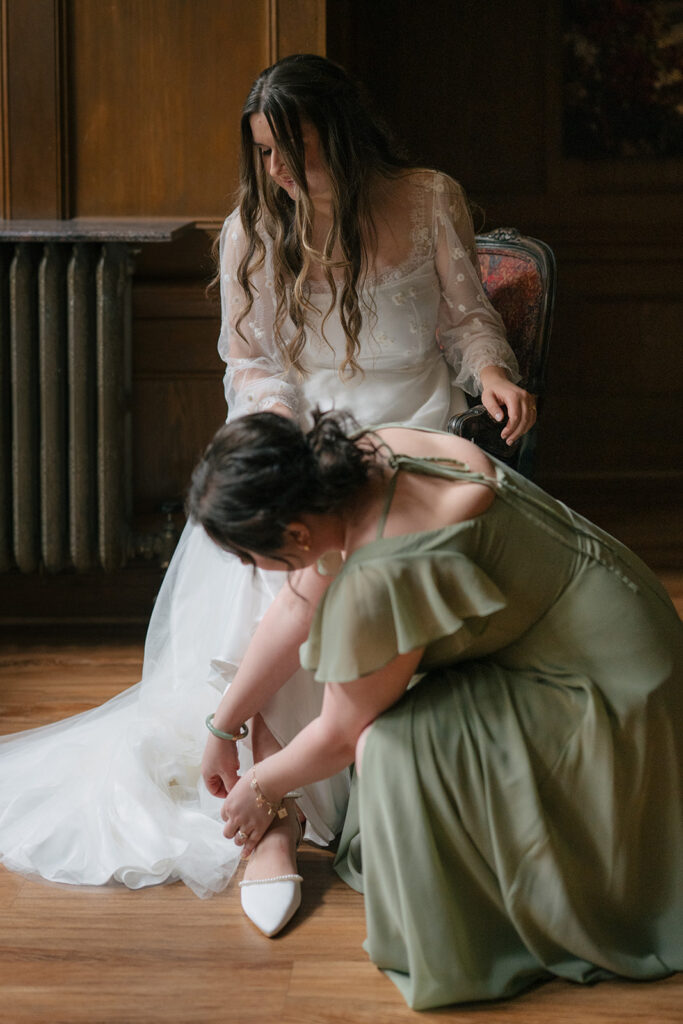 Bride seated as a bridesmaid secures her shoes before the ceremony