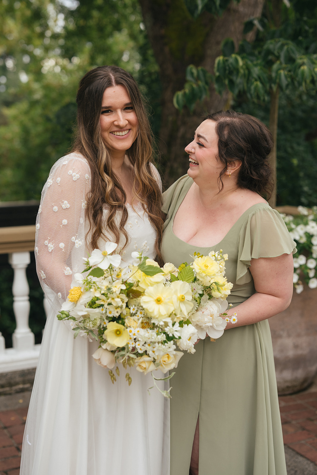 Bride holding her bouquet while smiling beside a bridesmaid in a sage green dress.