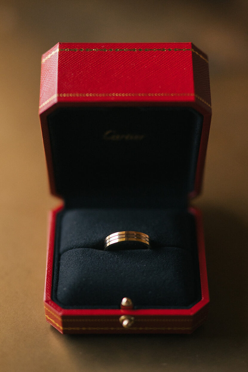 Gold wedding band inside a red ring box photographed in soft window light.
