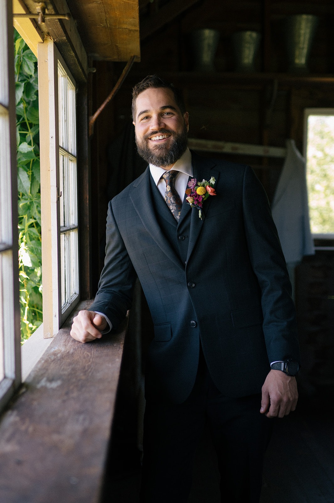 Groom leans against a window sill inside a rustic barn room, smiling