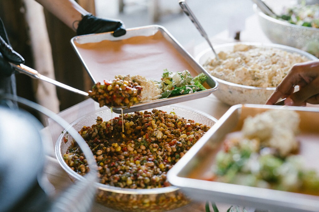 Catering staff spoon fresh bean salad into trays alongside creamy coleslaw