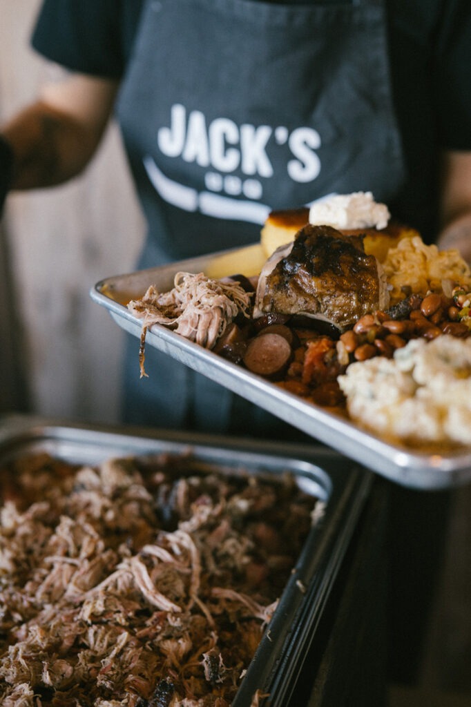 A caterer in a Jack's BBQ apron loads a tray piled with pulled pork and sides